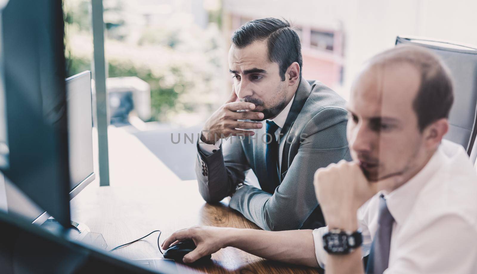 Image of two thoughtful businessmen looking at data on computer screens, solving business issue at business meeting in moder office.