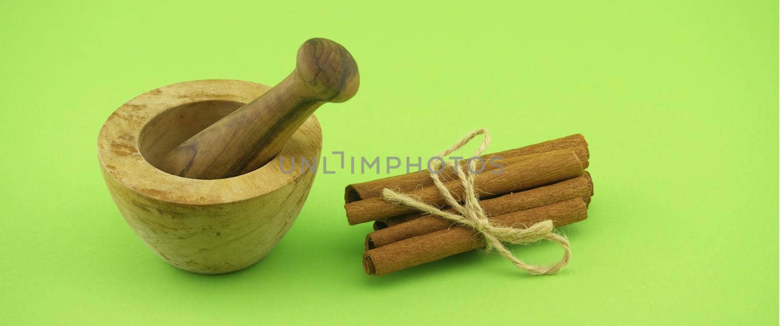 Wooden mortar and pestle near cinnamon sticks bundle tied with jute string over green background and free copy space for text