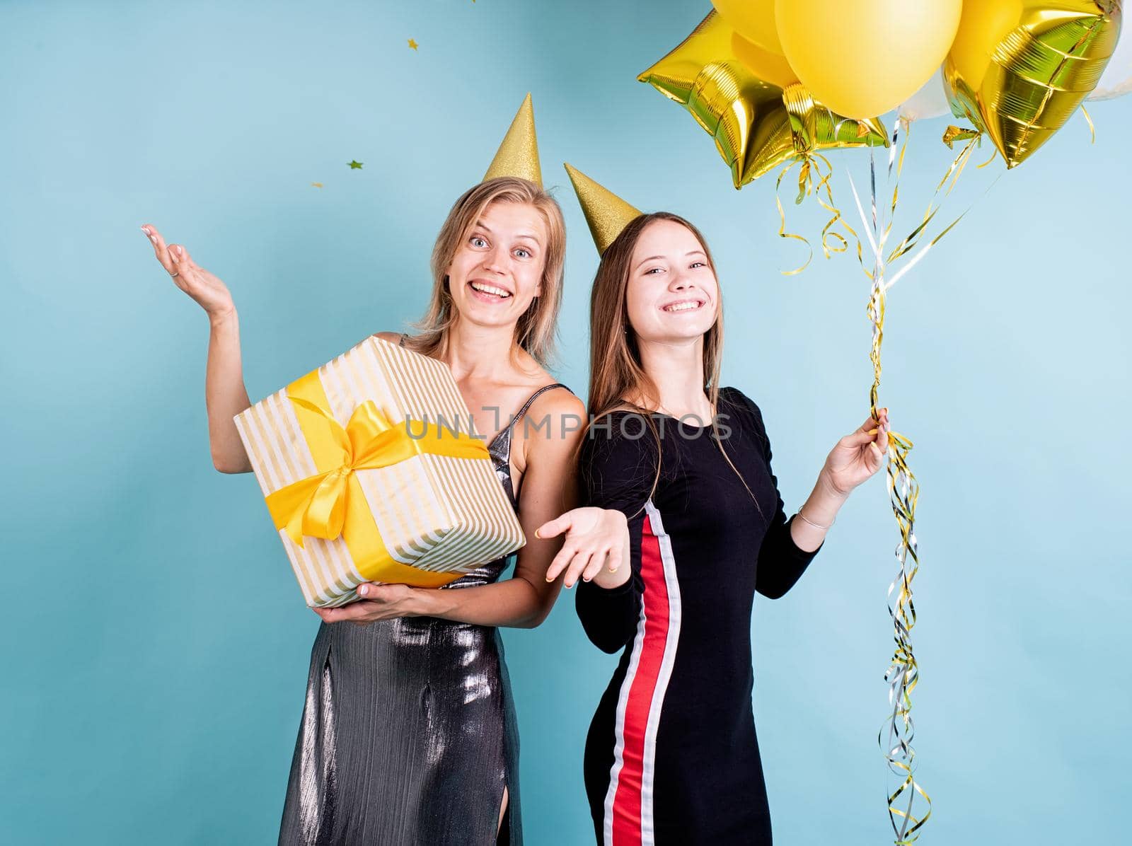 Two young women in birthday hats holding balloons celebrating birthday over blue background by Desperada