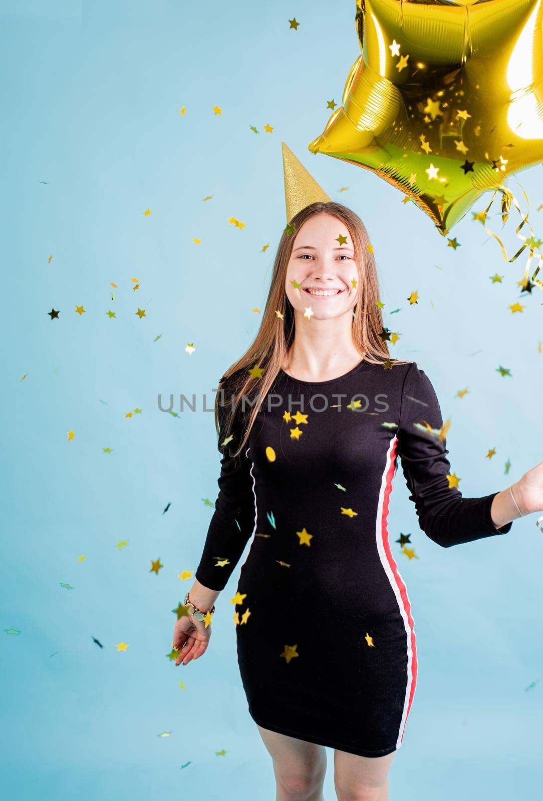 Birthday party. Teenager girl wearing golden birthday hat blowing confetti over blue background