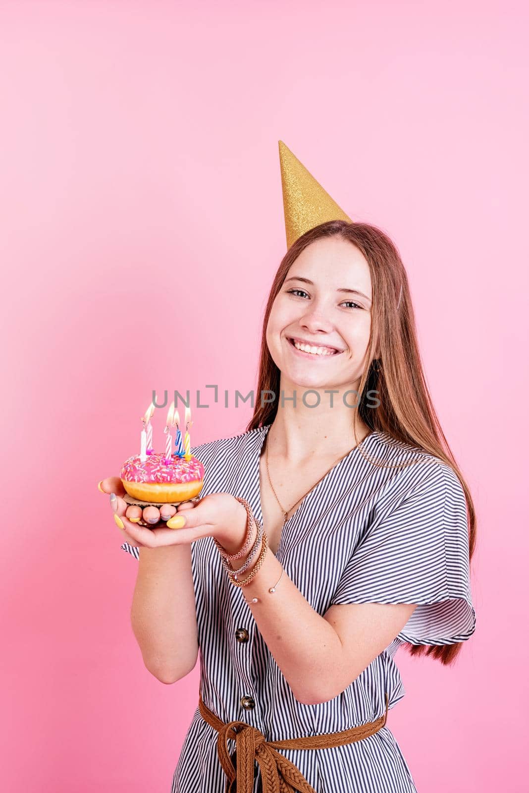 Birthday party. Teenager girl in golden birthday holding donut with candle, making a wish over pink background