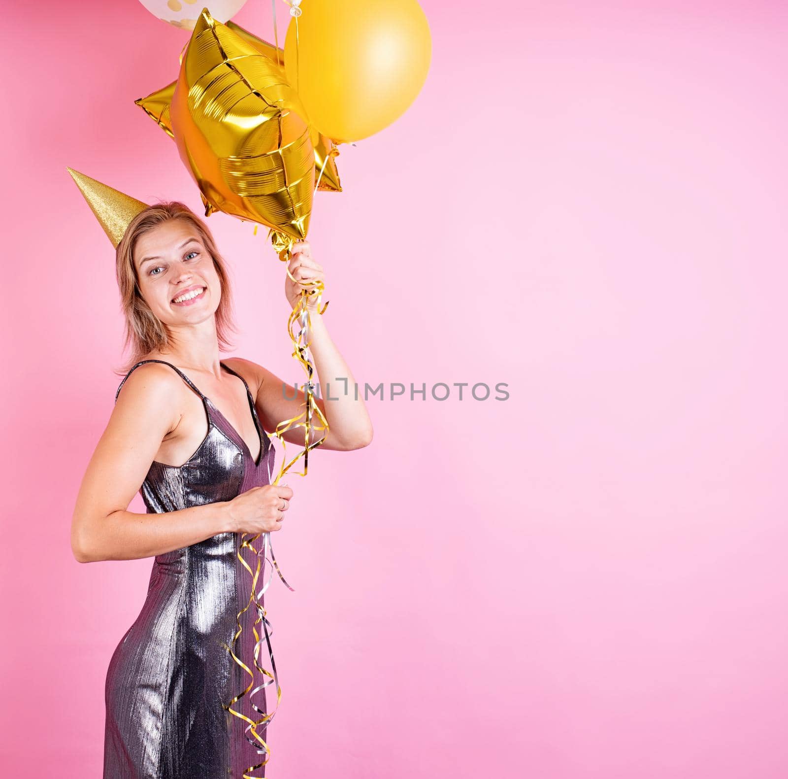 Birthday party. Young blond smiling woman wearing birthday hat holding golden balloons celebrating birthday on pink background with copy space