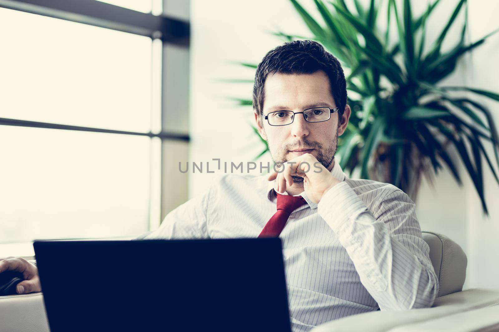 Portrait of successful young businessman wearing glasses in bright modern office with his laptop computer . Business and entrepreneurship concept.