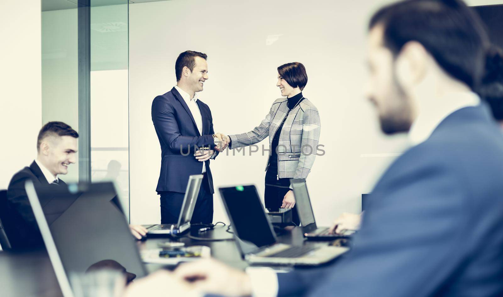 Sealing a deal. Business people shaking hands, finishing up meeting in corporate office. Businessman working on laptop in foreground. Business and entrepreneurship concept.