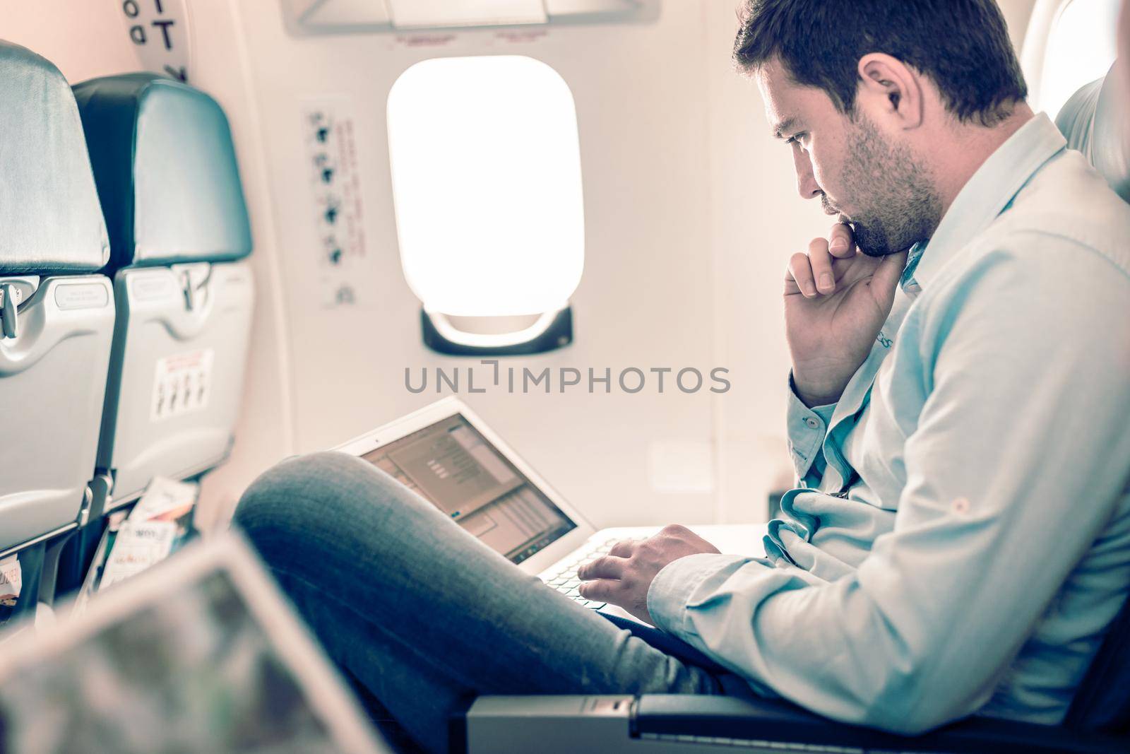 Casually dressed middle aged man working on laptop in aircraft cabin during his business travel. Shallow depth of field photo with focus on businessman eye.