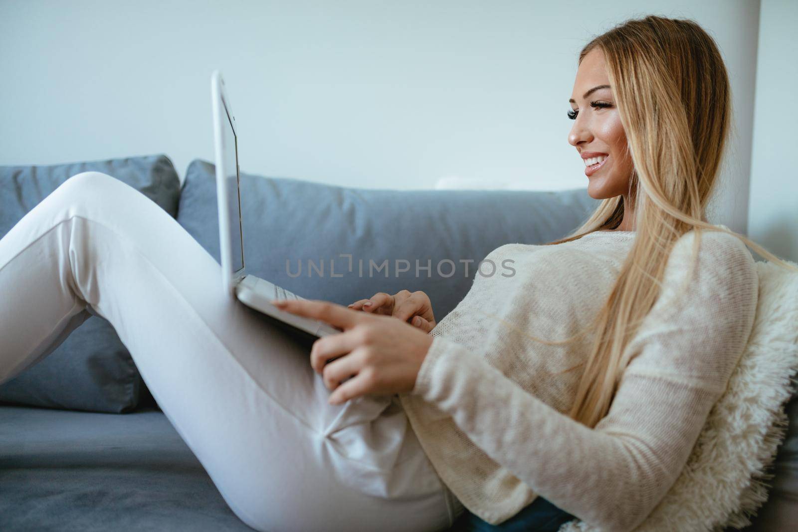 Attractive smiling girl sitting on a bed and using laptop. She is surfing on the net and checking social media.