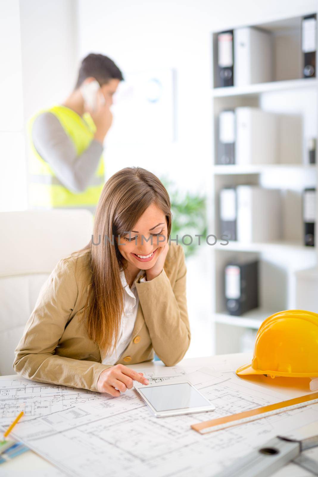 Smiling young woman constructor analyzing blueprint on tablet at desk in office.