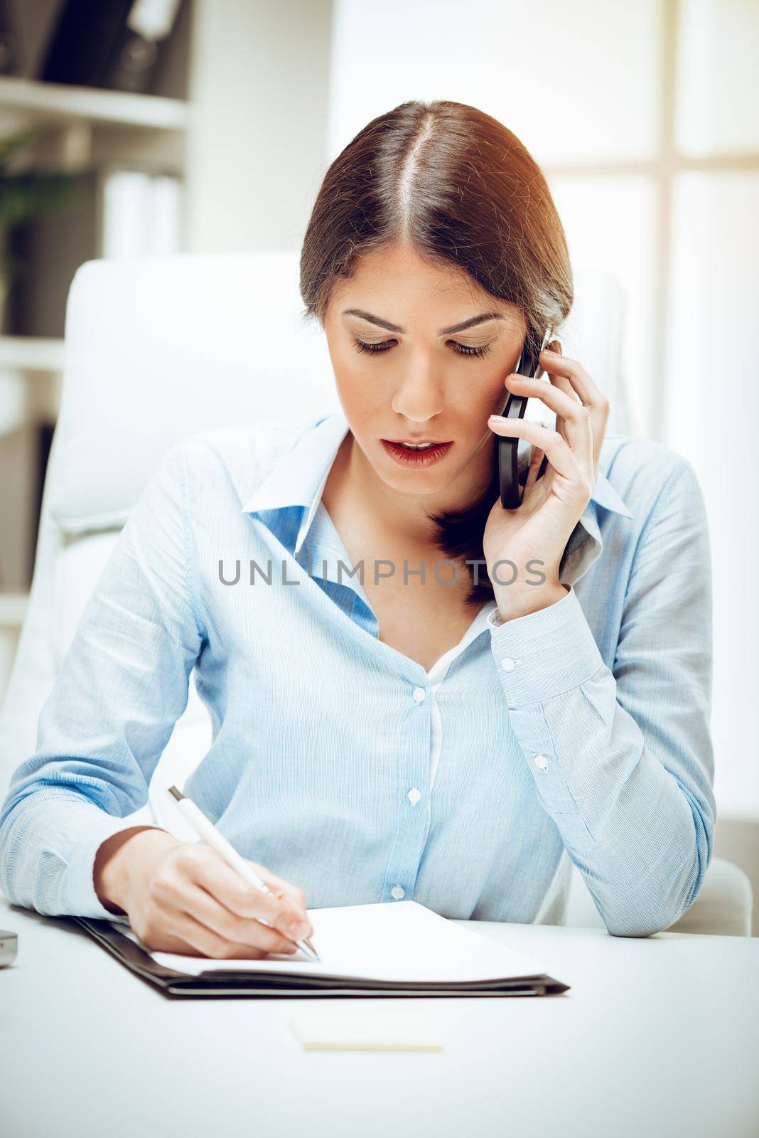 Successful young woman working at her desk in the office. She is using smart phone and analyzing business report. 