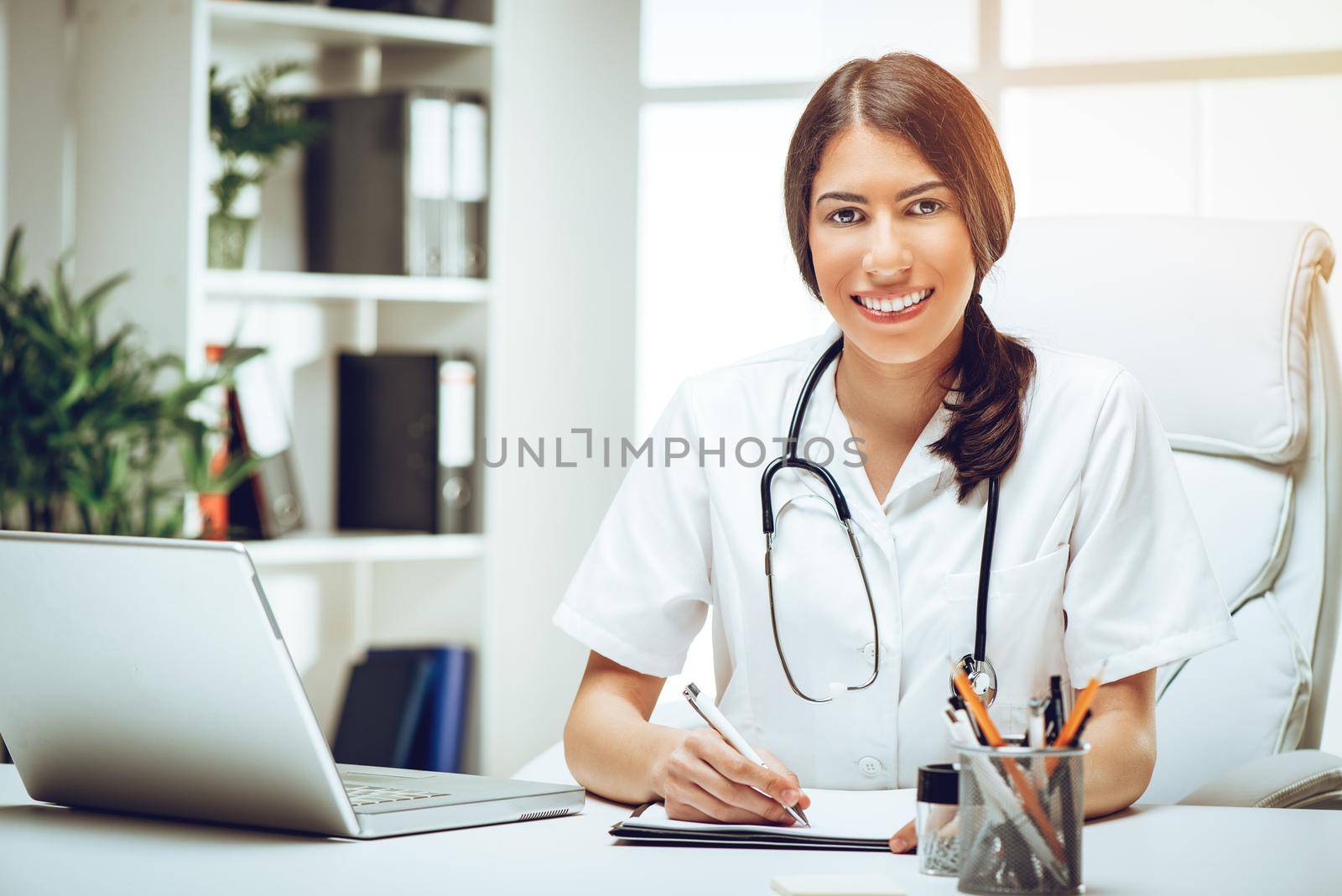 Portrait of a beautiful young female doctor sitting in her consulting room, smiling and looking at camera.