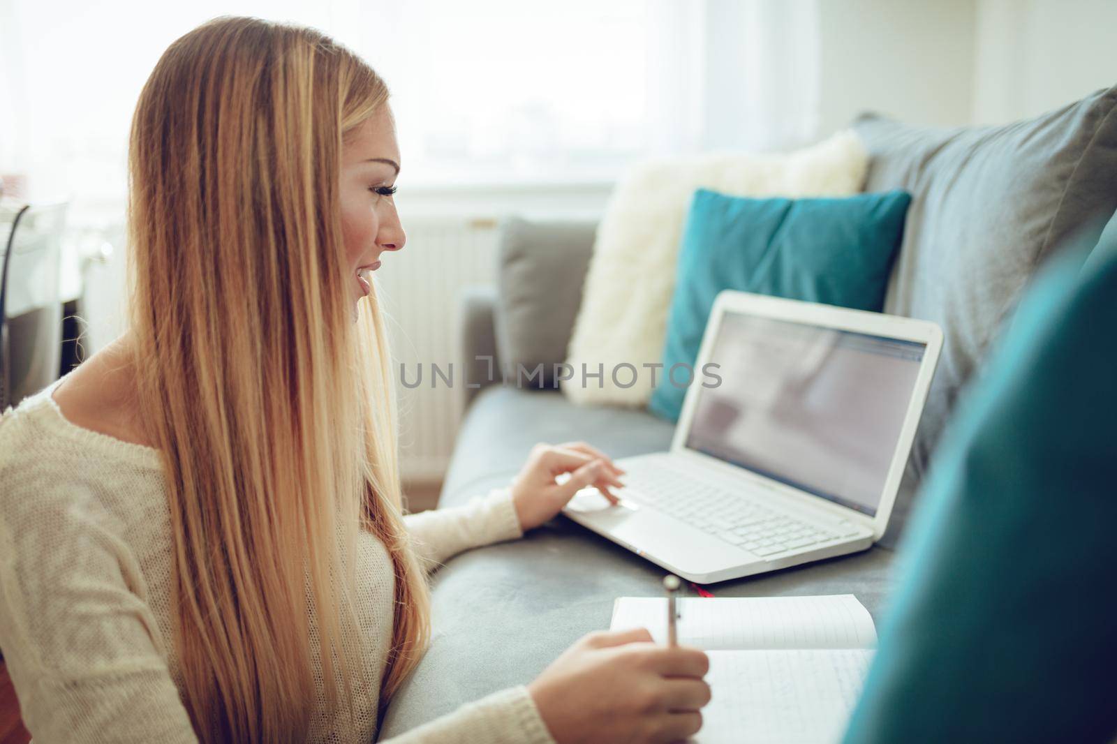 Beautiful young woman studying and working on her laptop and writing at the notebook, sitting at the floor beside the sofa at home.