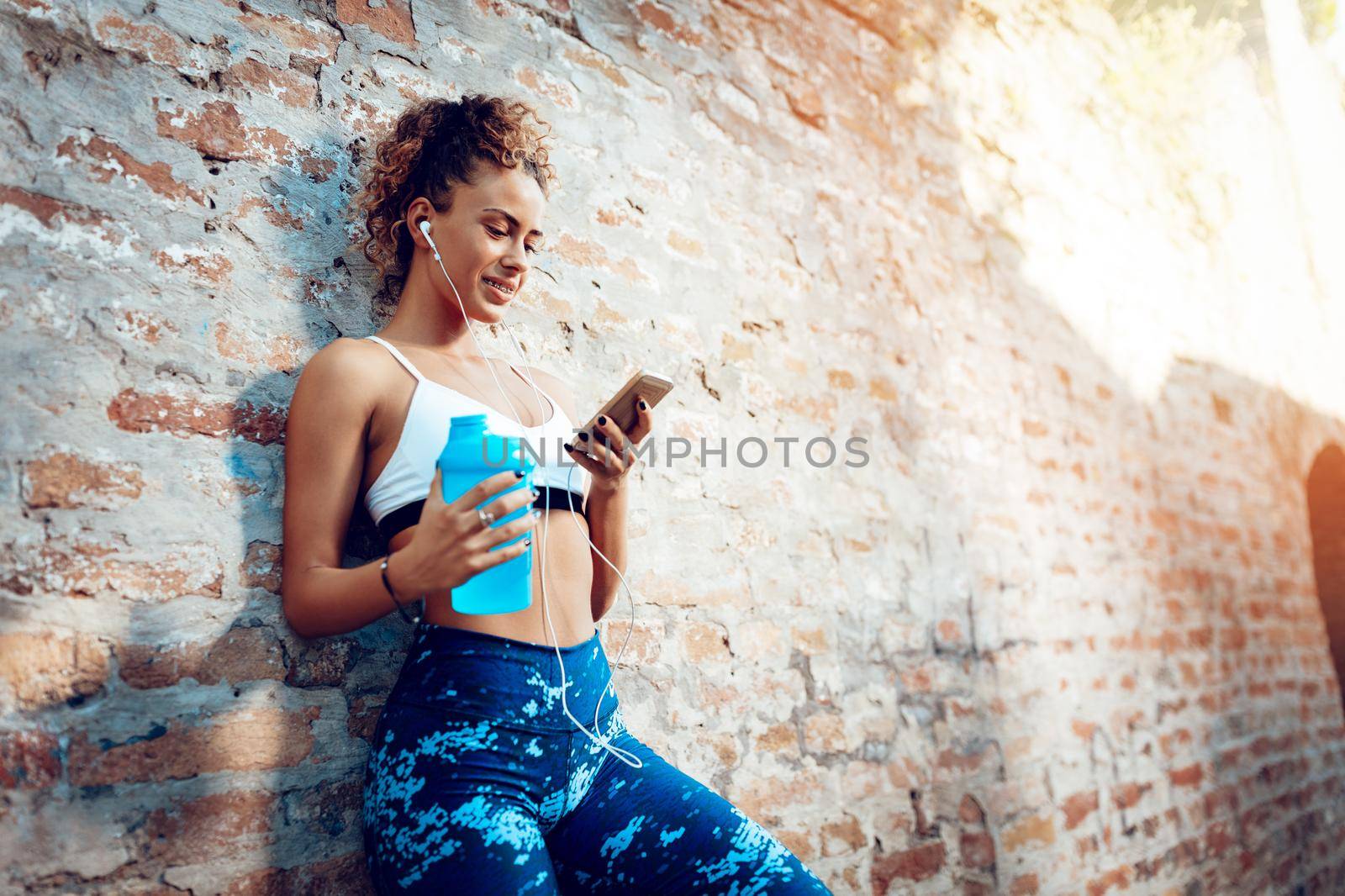 Young fitness smiling woman with headphones taking a break after running. She is standing up against the wall and using mobile phone.