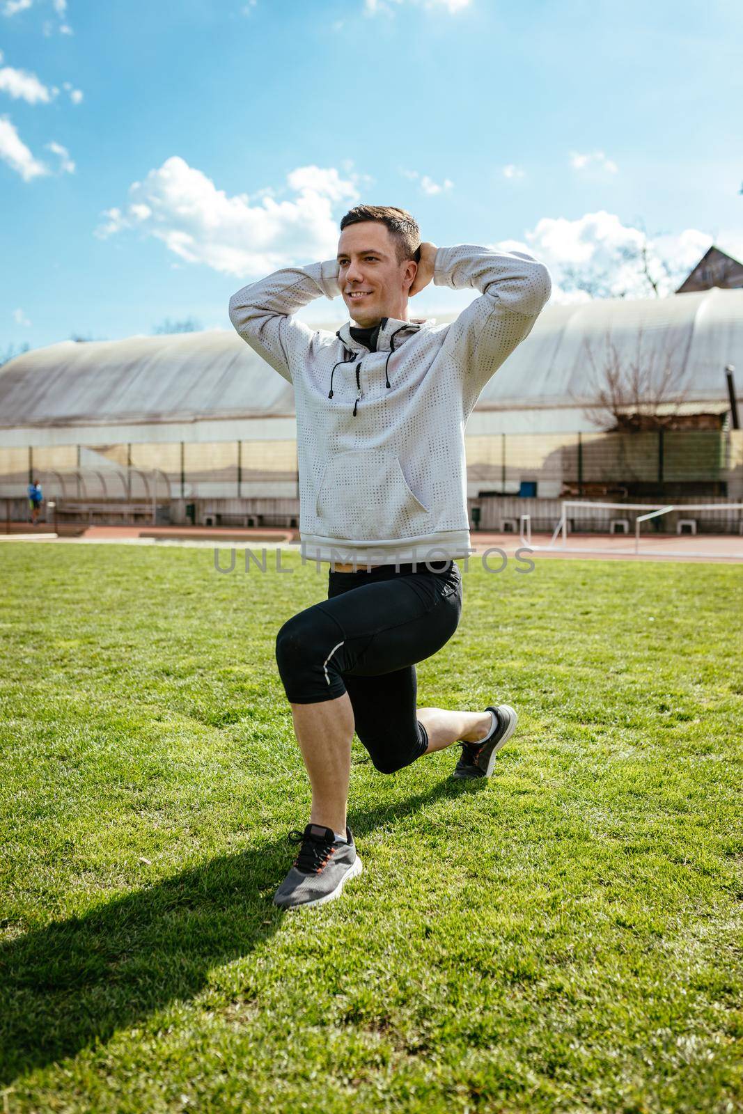 Active young man stretching and doing one leg crouching exercises in the public place on empty stadium. 