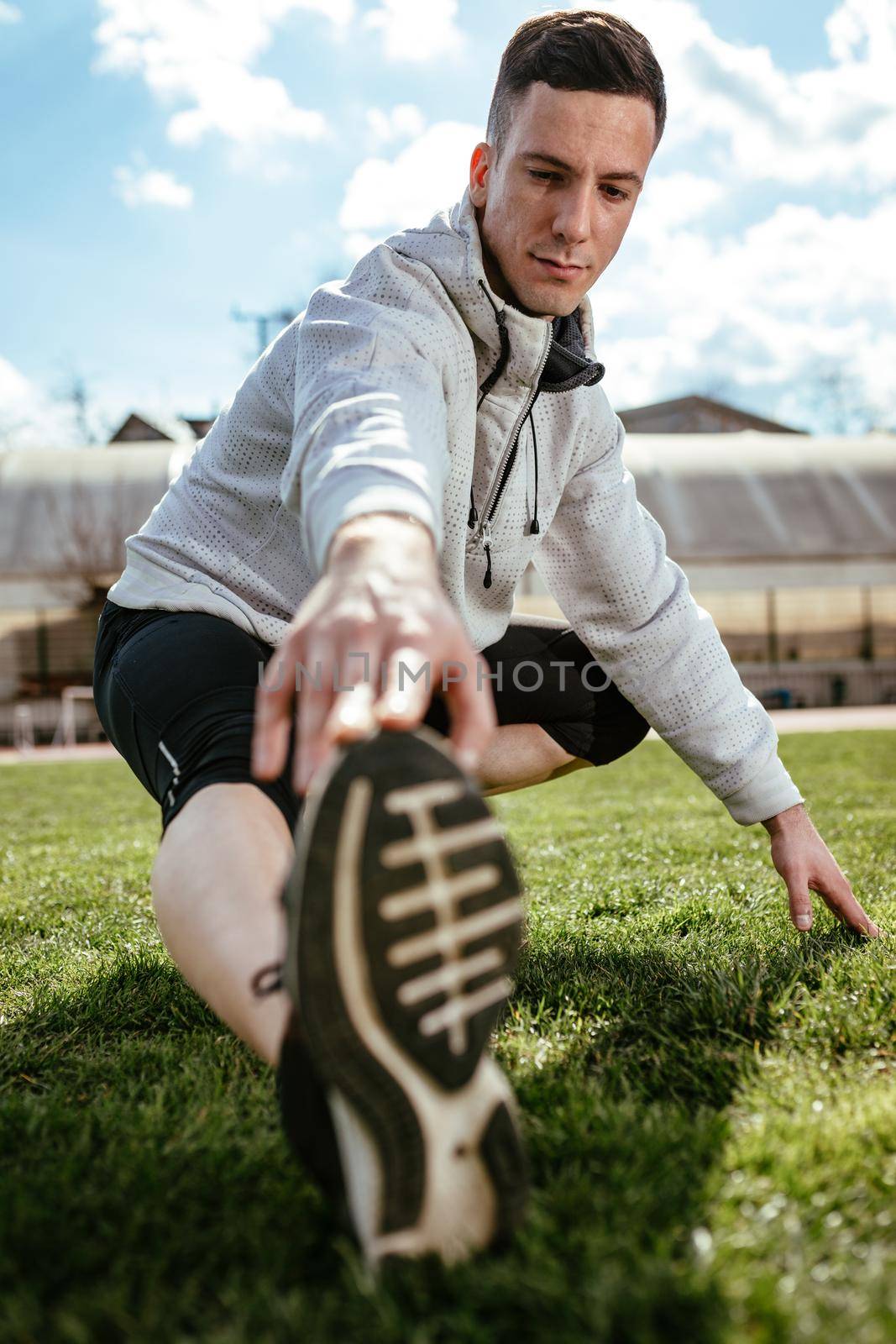 Active young man stretching legs and doing exercises in the public place on empty stadium. 