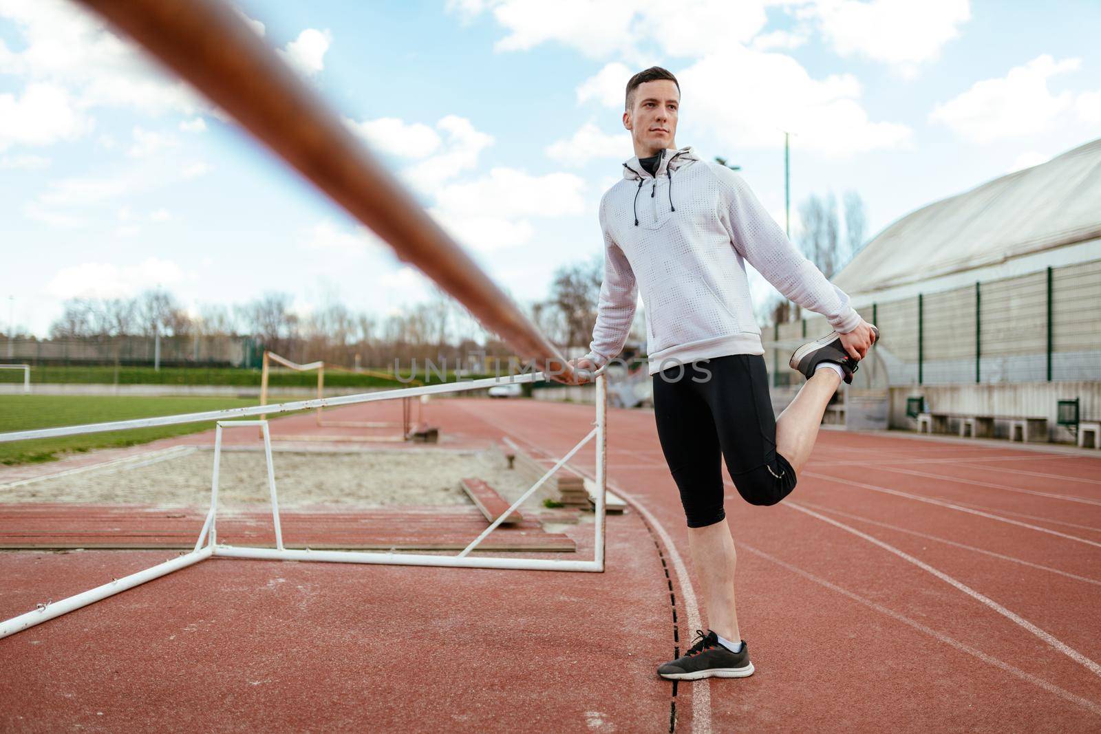 Active young man stretching and doing exercises preparing to run over tartan track on the stadium. 