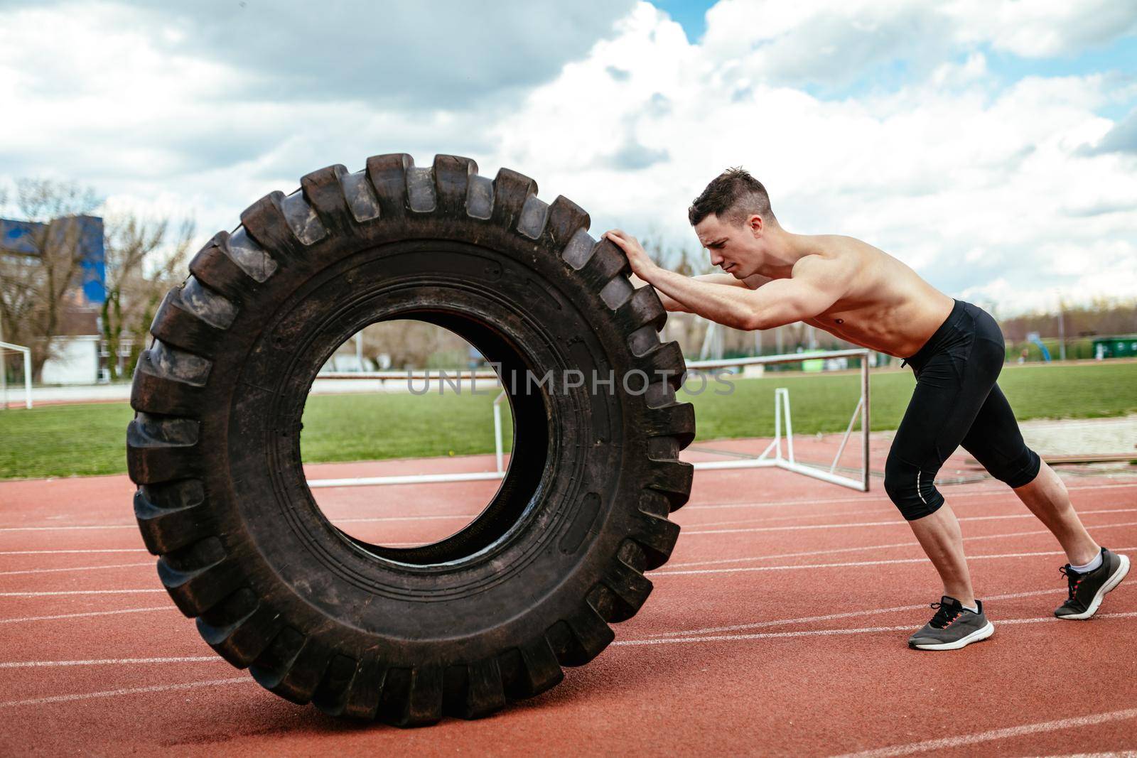 Young muscular man flipping a huge tire for training muscles at the tartan track on the stadium.