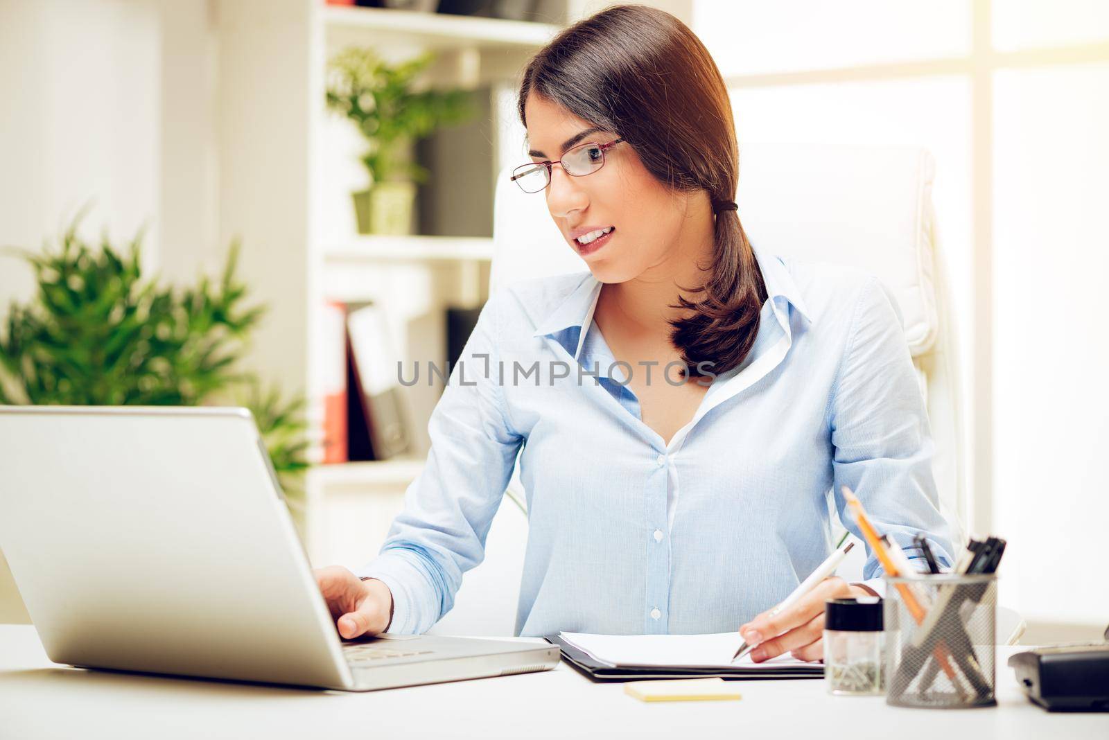 Successful young woman working at her desk in the office. She is using laptop and analyzing business report. 