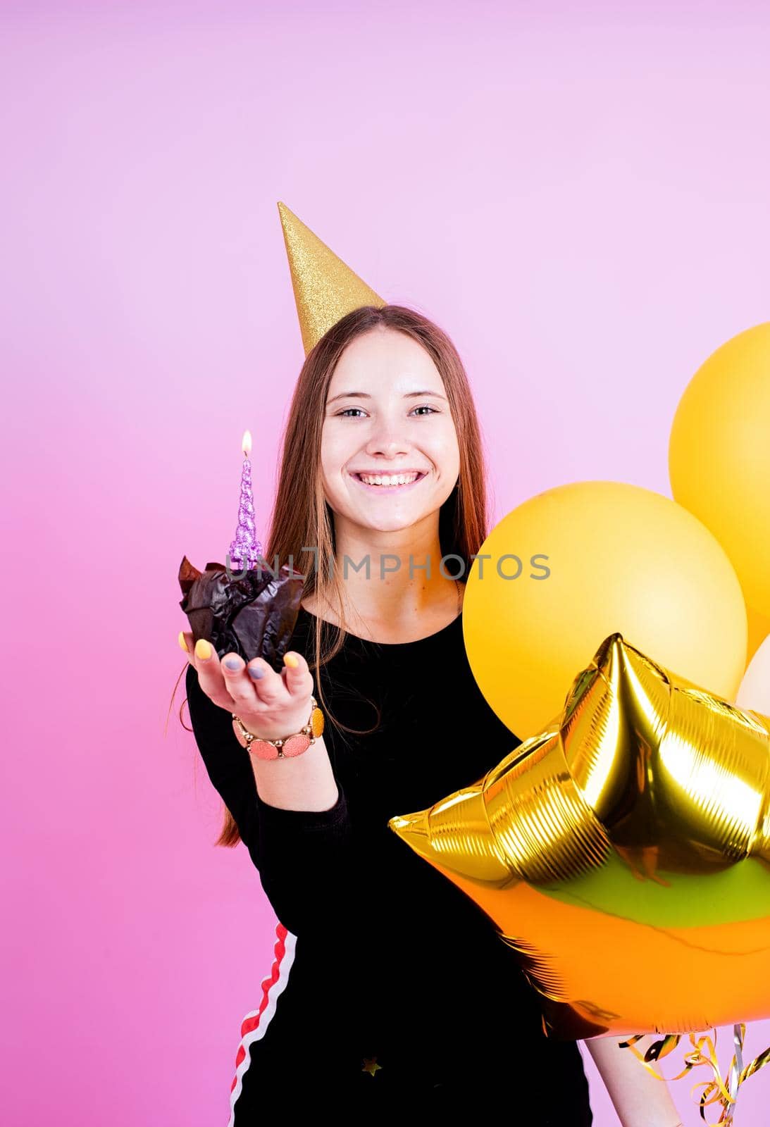 Birthday party. Teenager girl in golden birthday holding muffin with a candles, making a wish over pink background