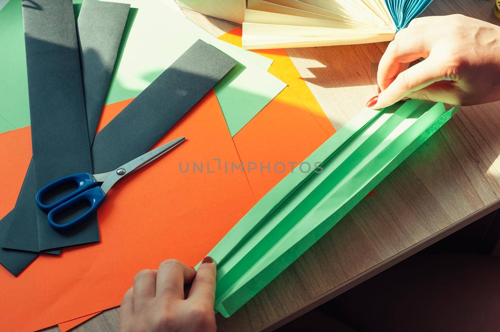 In the foreground, female hands are preparing blanks for an origami fan, sheets of colored paper, scissors on a wooden table. Several blanks for a fan. Template for design, advertising or text.