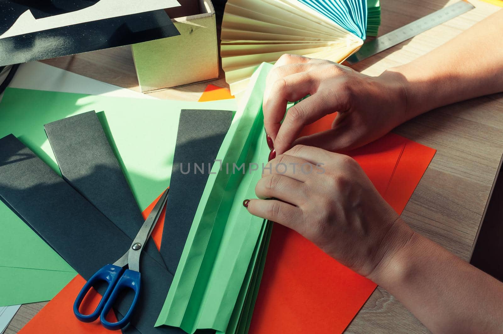 In the foreground, female hands are preparing blanks for an origami fan, sheets of colored paper, scissors on a wooden table. Several blanks for a fan. Template for design, advertising or text.