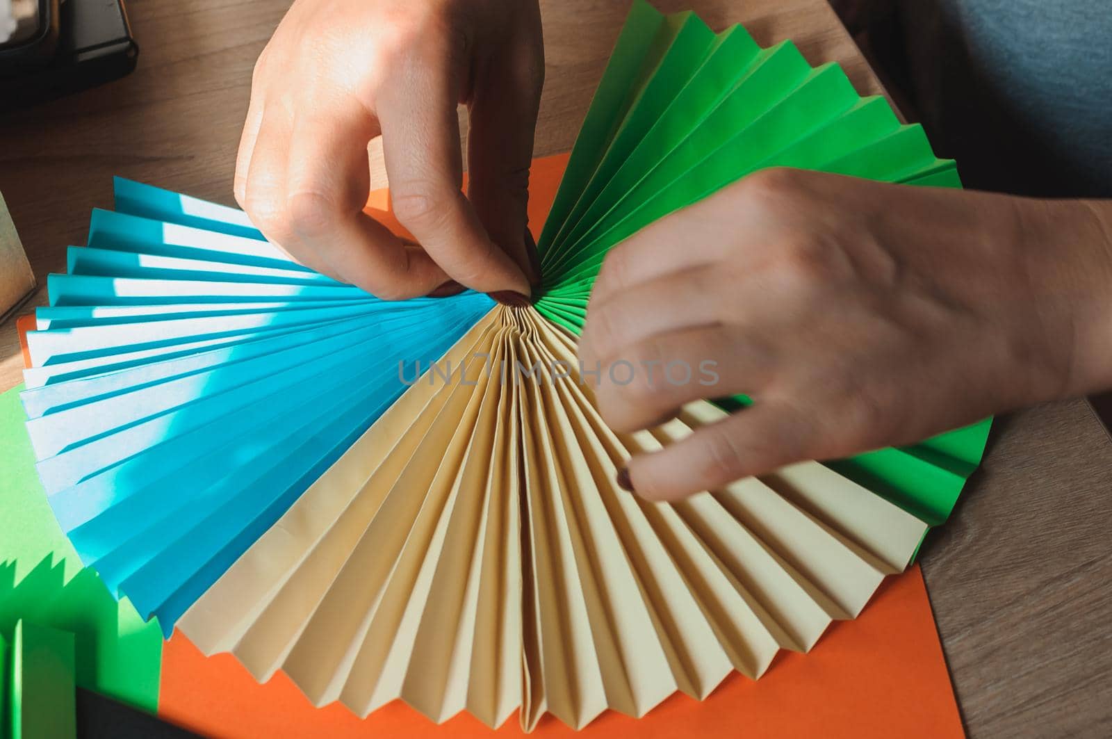 In the foreground female hands glue origami fans, sheets of colored paper, scissors on a wooden table. Several blanks for the fan. Template for design, advertising or text.