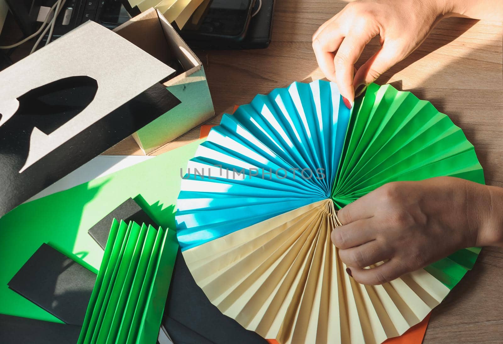 In the foreground female hands glue origami fans, sheets of colored paper, scissors on a wooden table. Several blanks for the fan. Template for design, advertising or text.