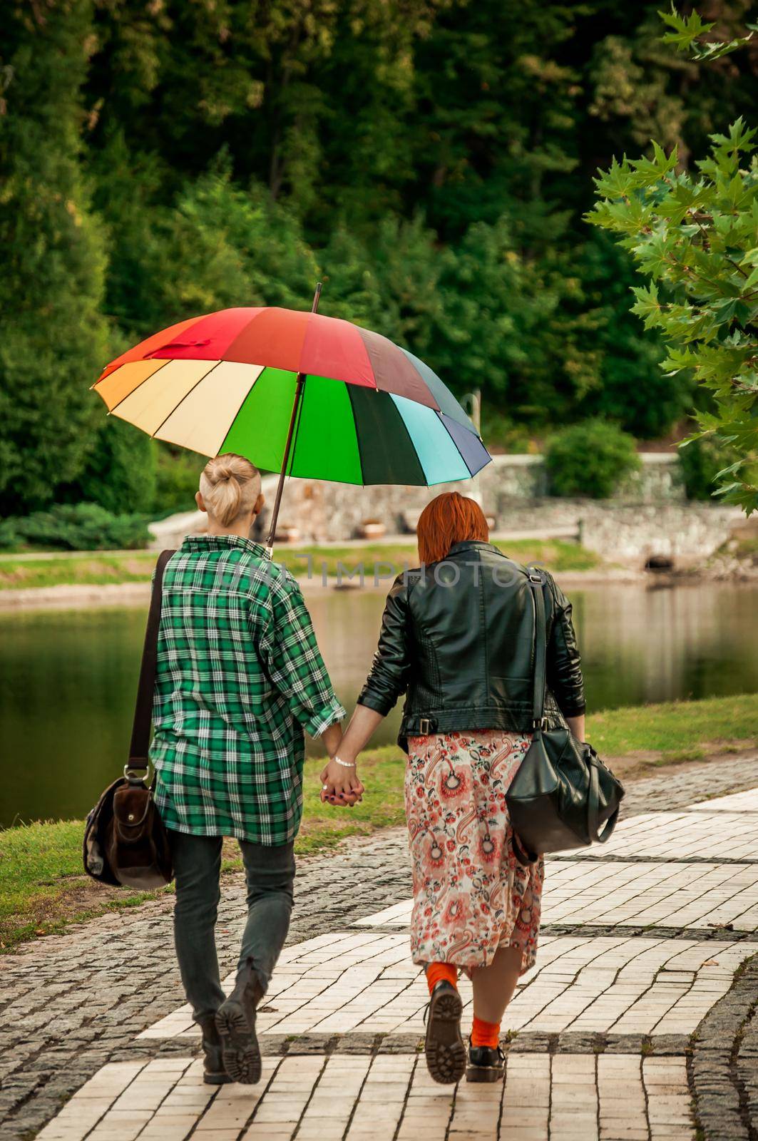 Lesbian Couple holding hands walking in the autumn park in the rain with a rainbow umbrella. The concept of free love and walks in the park. LGBT community by Alla_Morozova93