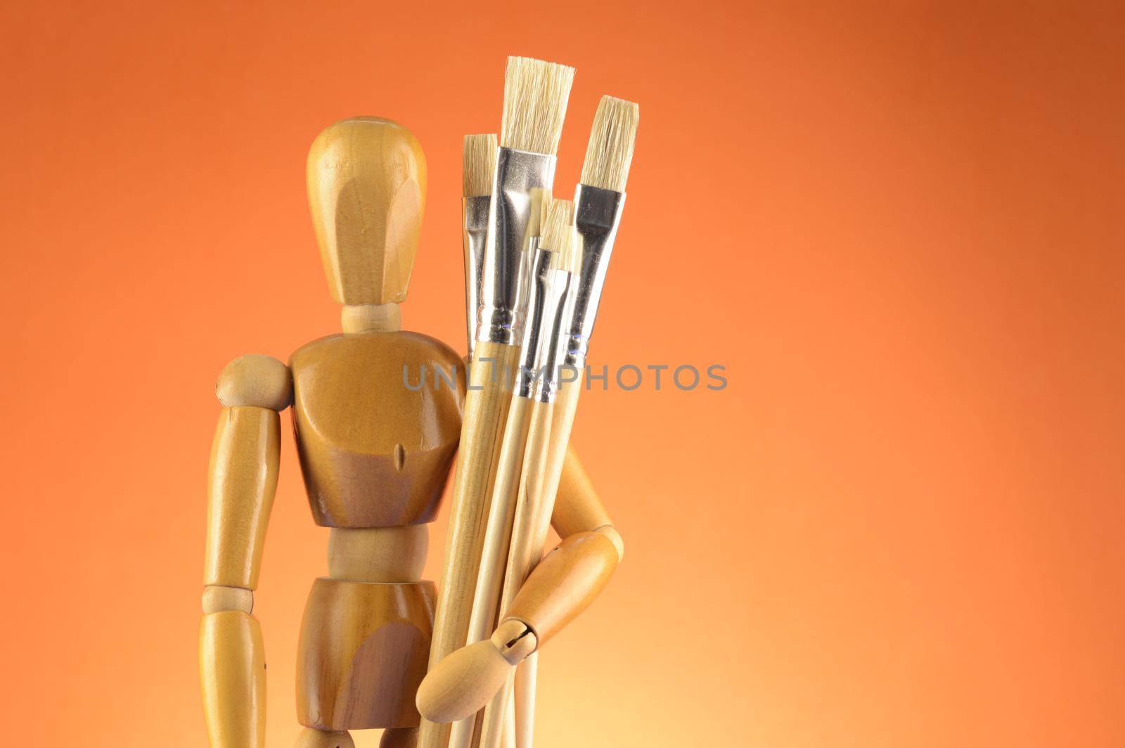 An Artists wooden Dummy Mannequin stands with some dry paintbrushes over an orange background.