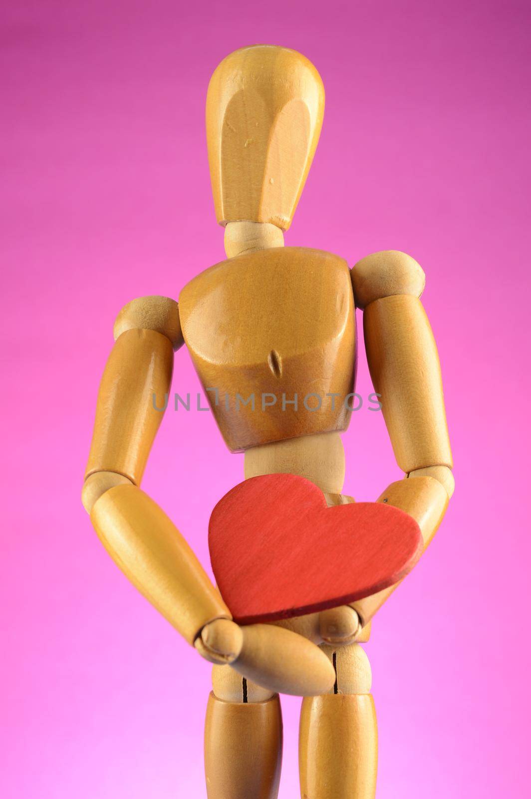 An Artist wooden Dummy Mannequin holds a red heart closely while standing over a pink background.