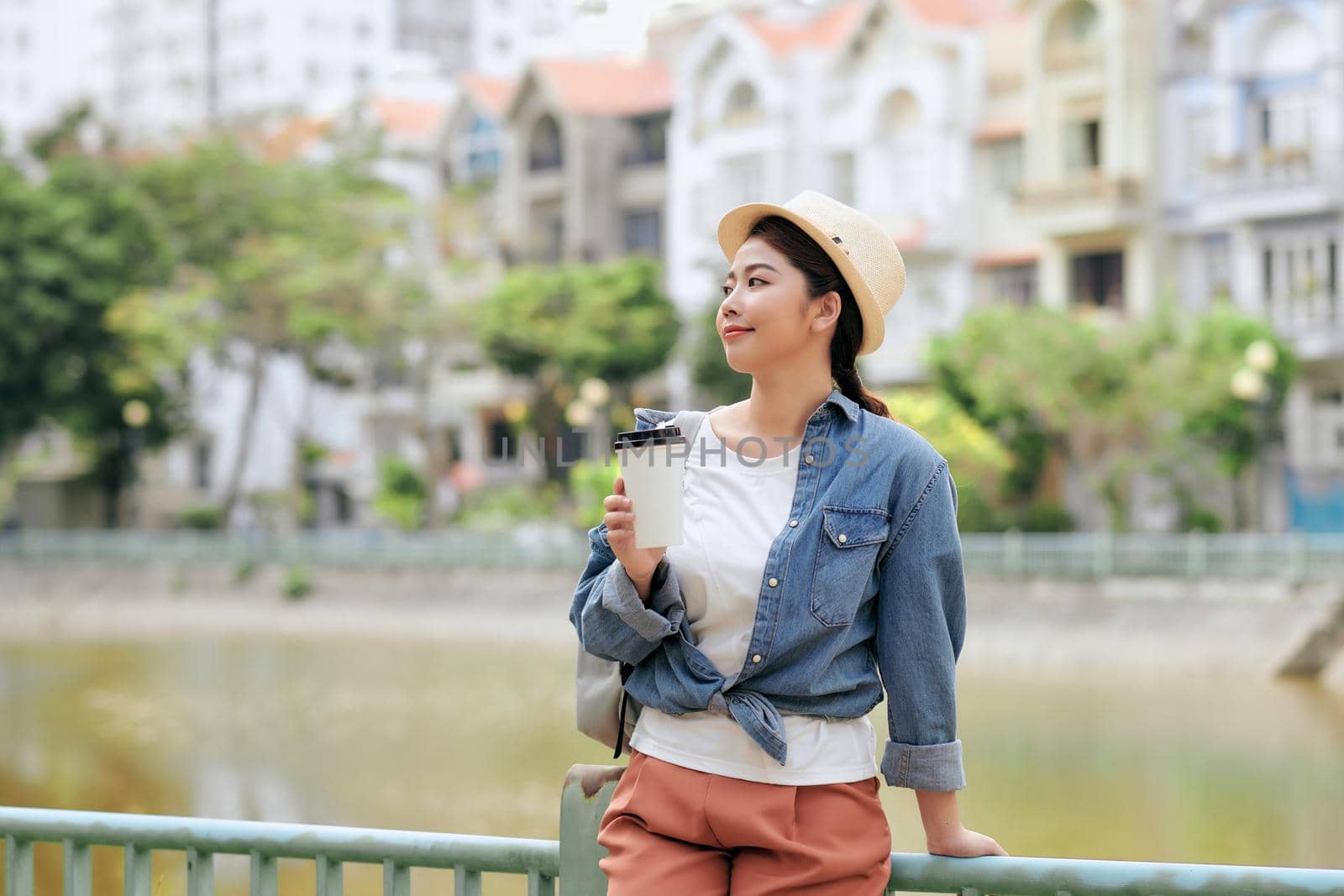 Pleased woman in casual wear holding in hand paper cup with hot drink while standing in city street at daytime