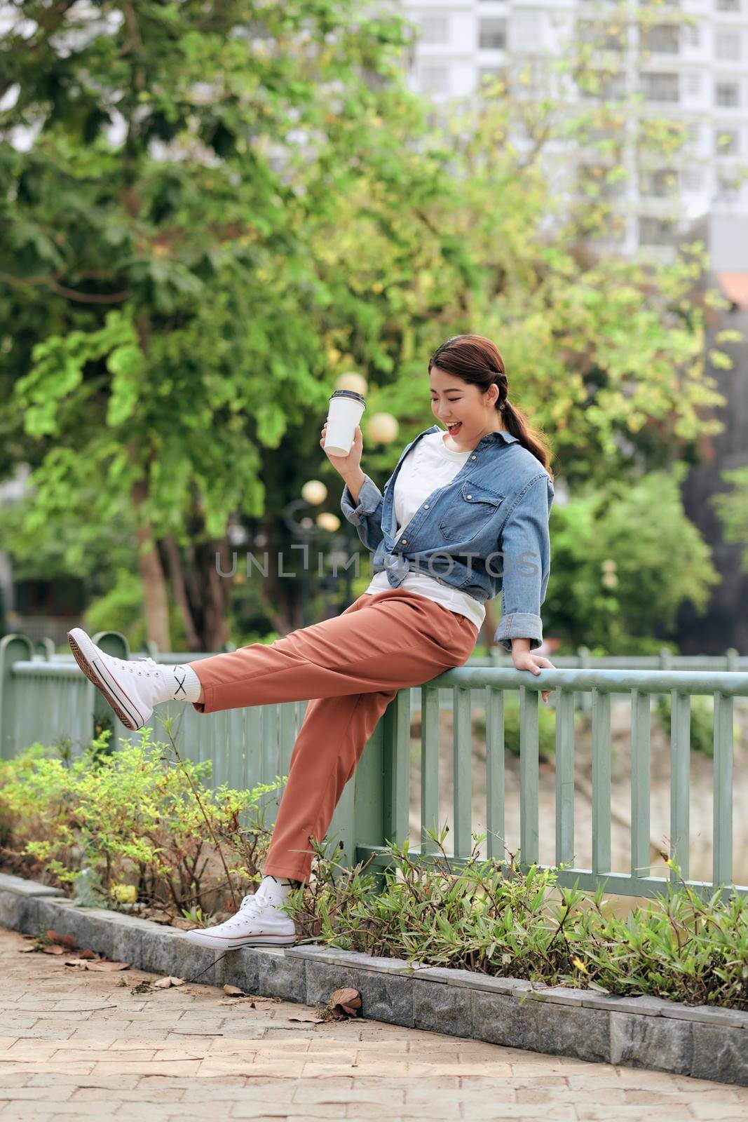 Attractive cheerful trendy girl in stylish denim jacket drinks takeaway coffee or tea outdoors