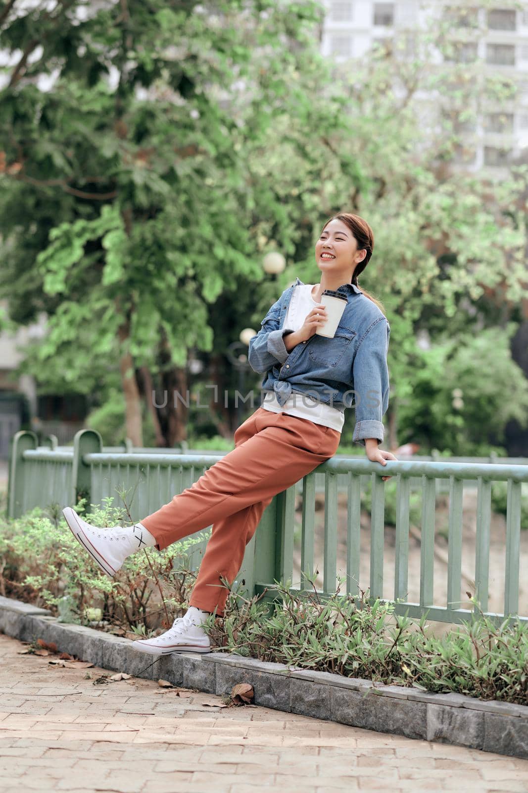 Happy young beautiful woman holding coffee cup in hands and smiling while sitting on city street