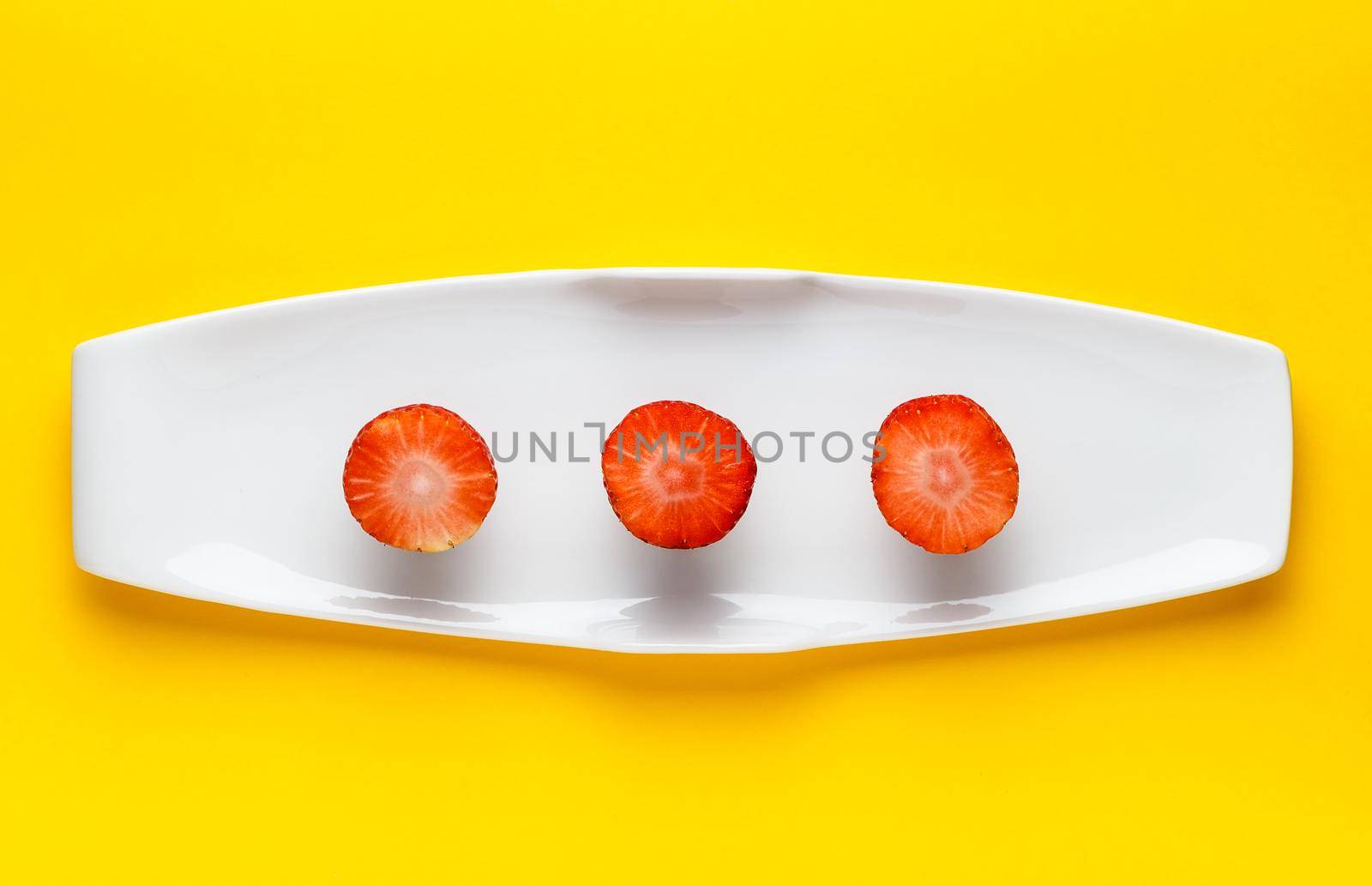 Three slices of fresh strawberry on a white elongated plate on yellow background. Horizontal image seen from above.