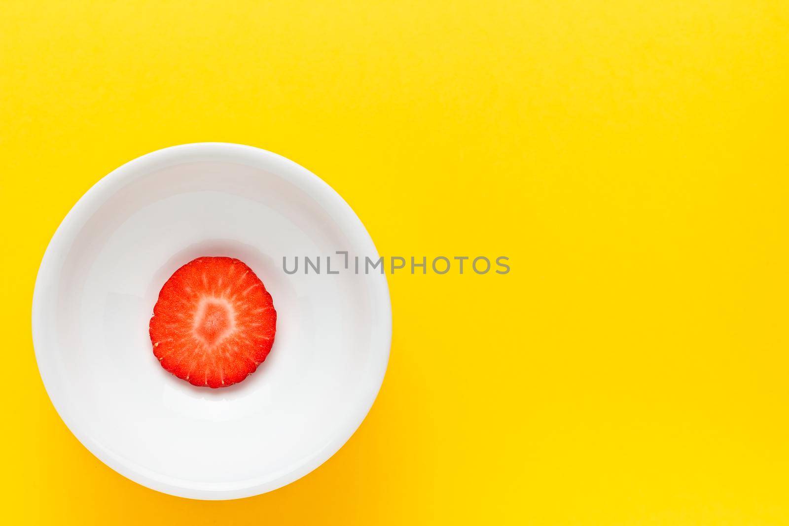 Slice of fresh strawberry on a round white plate on yellow background. Horizontal image seen from above.
