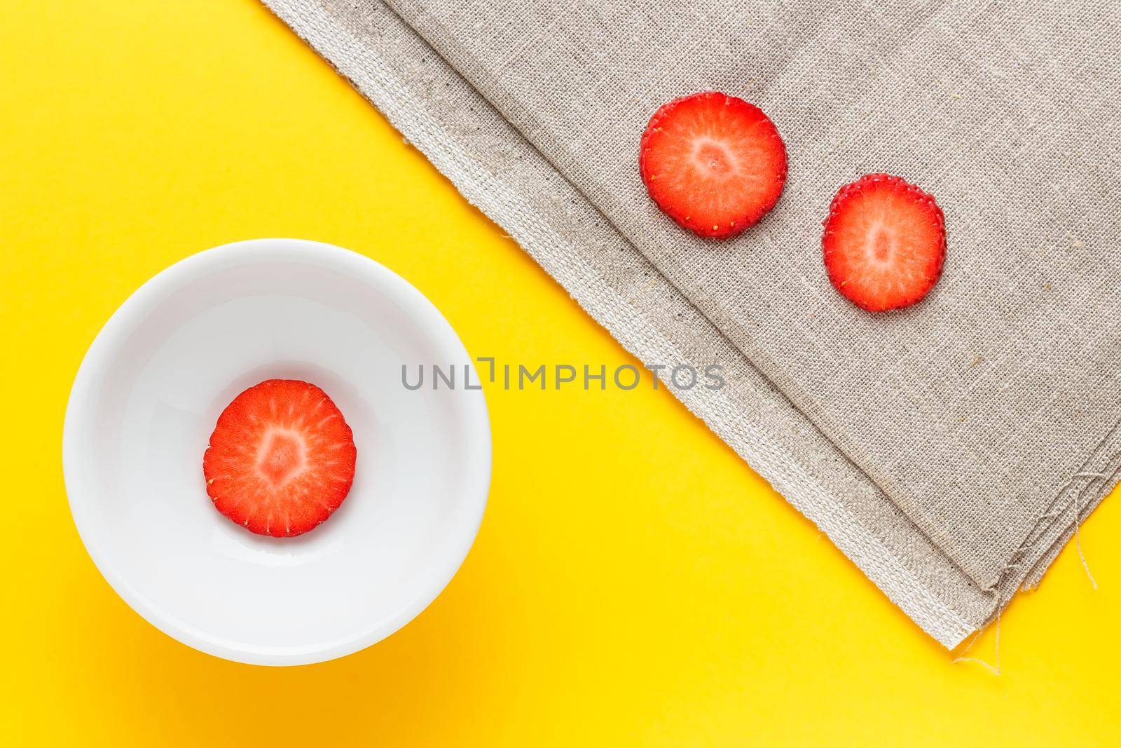 Three slices of fresh strawberries in a round white plate on yellow background and sackcloth. Horizontal image viewed from above.