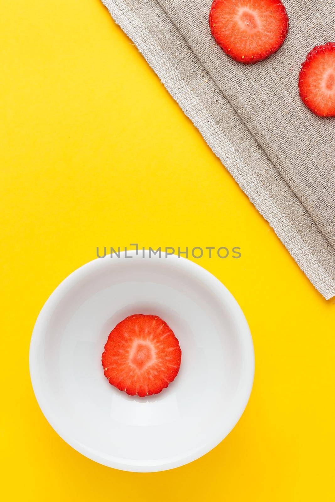 Three slices of fresh strawberries in a round white plate on yellow background and sackcloth. Vertical image viewed from above.