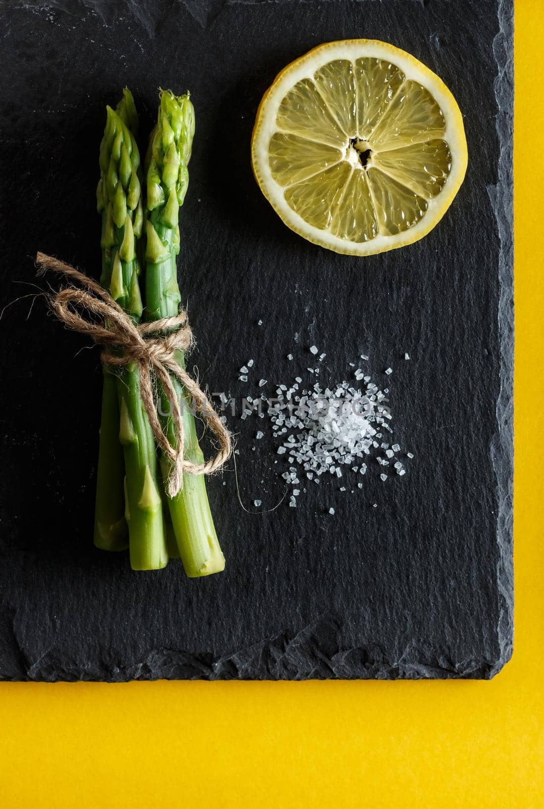 Asparagus. Bunch of fresh green asparagus with coarse salt and a slice of lemon on a slate plate on yellow background. Healthy vegetarian food. Vertical image
