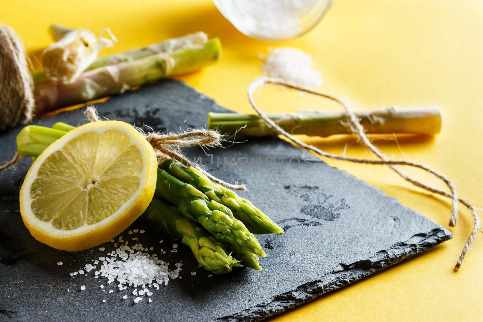 Asparagus. Bunch of fresh green asparagus close up with coarse salt and a slice of lemon on a slate plate on yellow background. Rope pieces and esparraggos cut to the bottom. Healthy vegetarian food. Horizontal image
