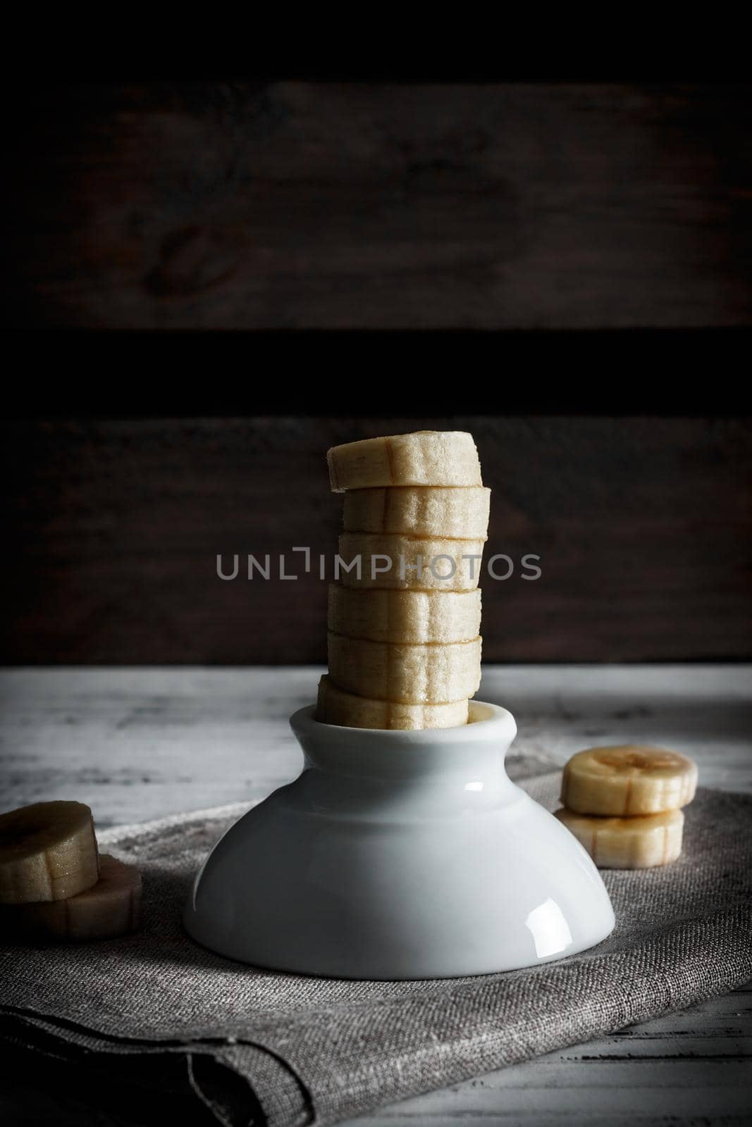 Delicious dessert of fresh sliced banana standing on a white bowl with slices around on a sackcloth and wooden background. Vertical image of dark mood style.