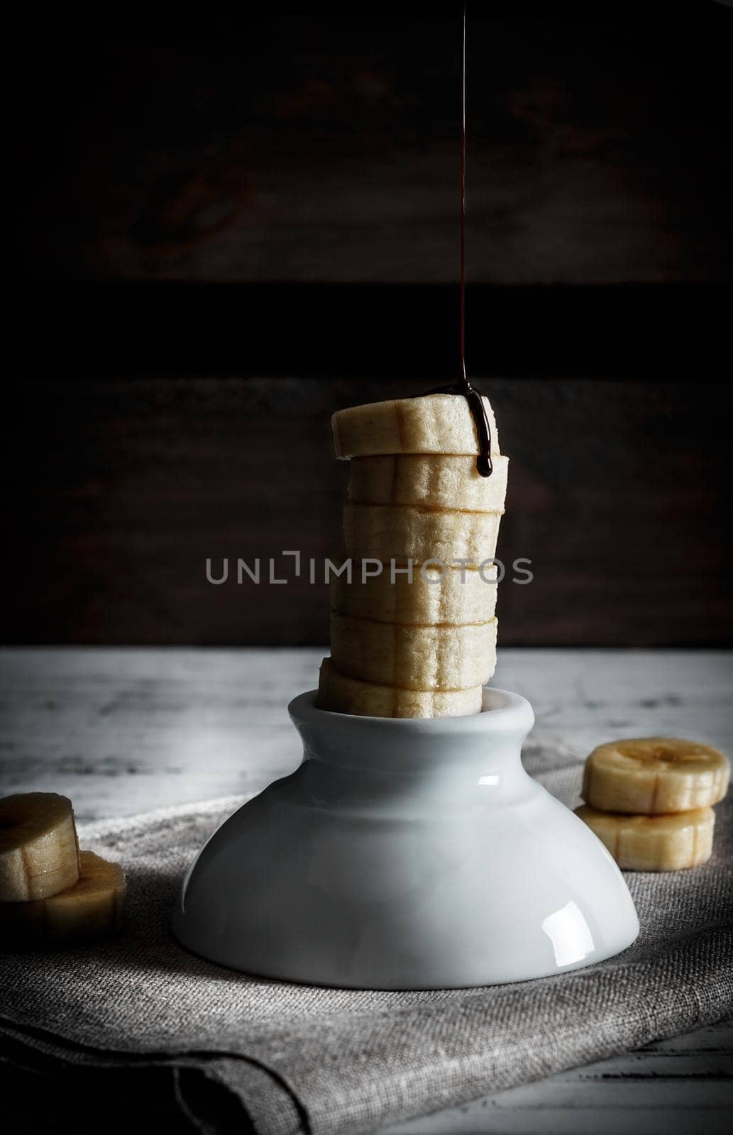 Delicious dessert of fresh banana slices standing with liquid chocolate on top on a white bowl with slices around on a sackcloth and a background of wooden boards. Vertical image of dark moody style.