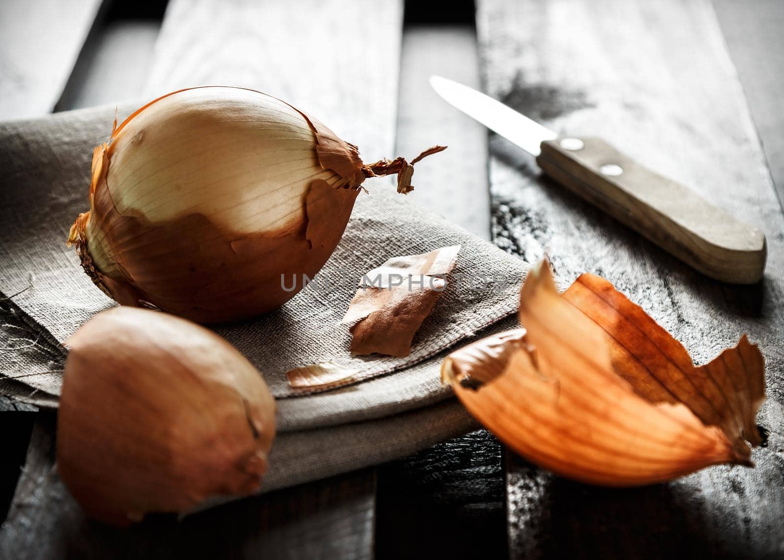 Still life of onion on a sackcloth with onion skins around and a knife in the background on wooden boards illuminated against the light. Dark moody style. Horizontal image.