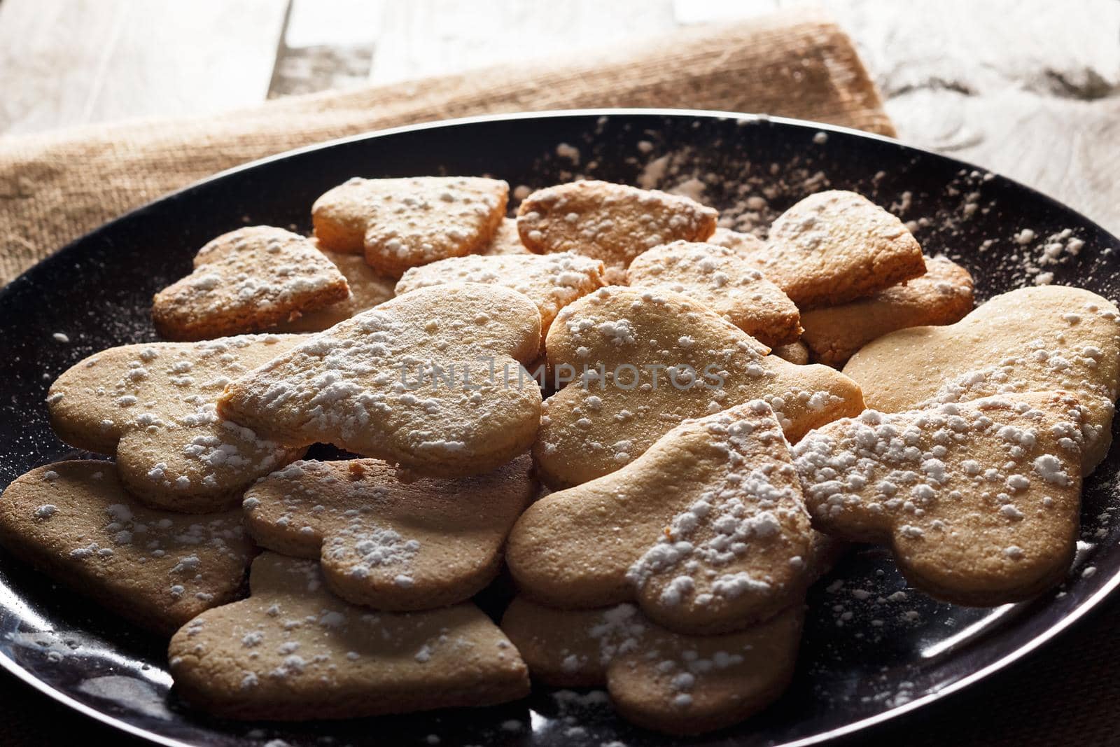 Delicious home-made heart-shaped cookies sprinkled with icing sugar on sackcloth and wooden boards. Horizontal image seen against backlight.