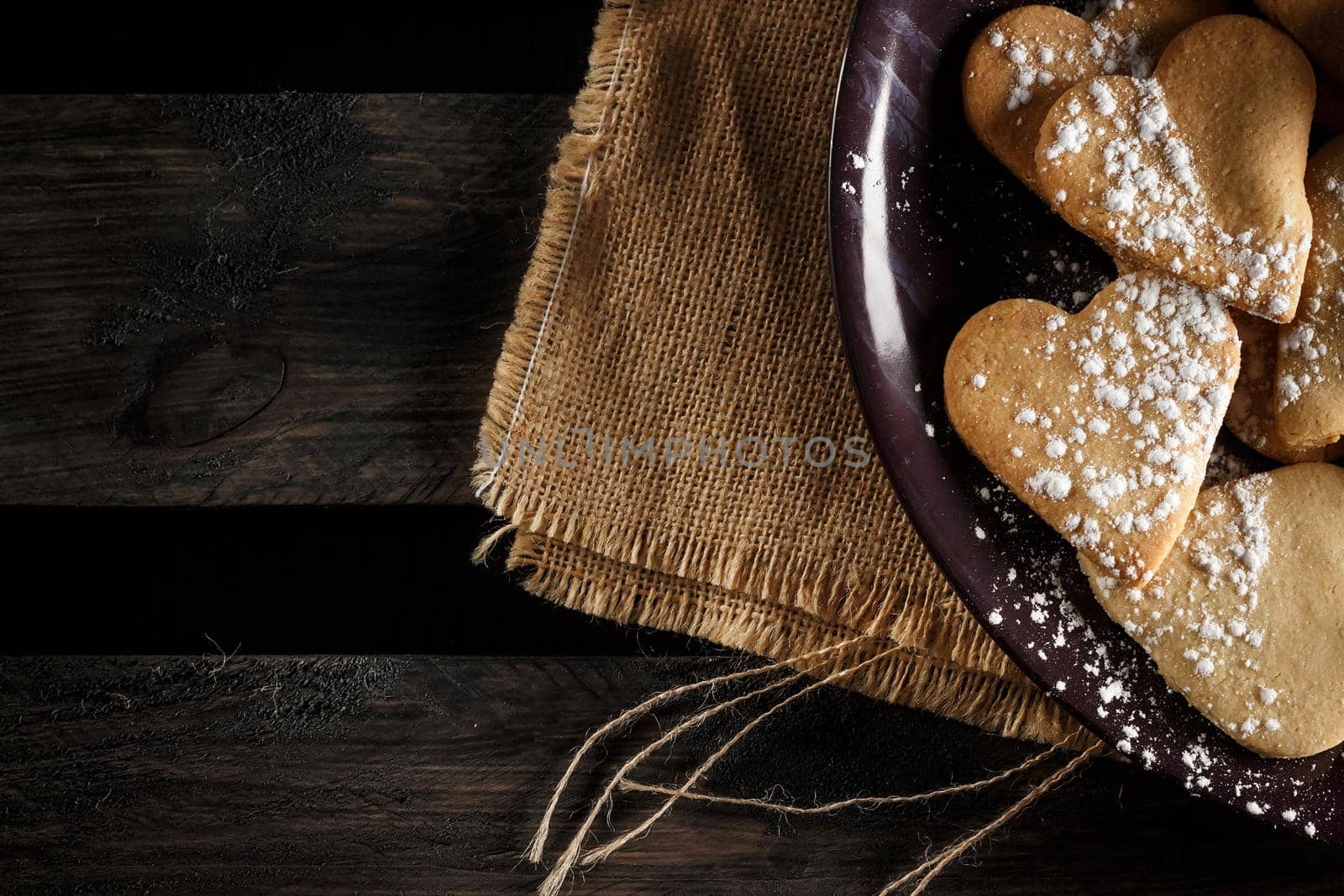 Delicious home-made heart-shaped cookies sprinkled with icing sugar on sackcloth and wooden boards. Horizontal image seen from above.