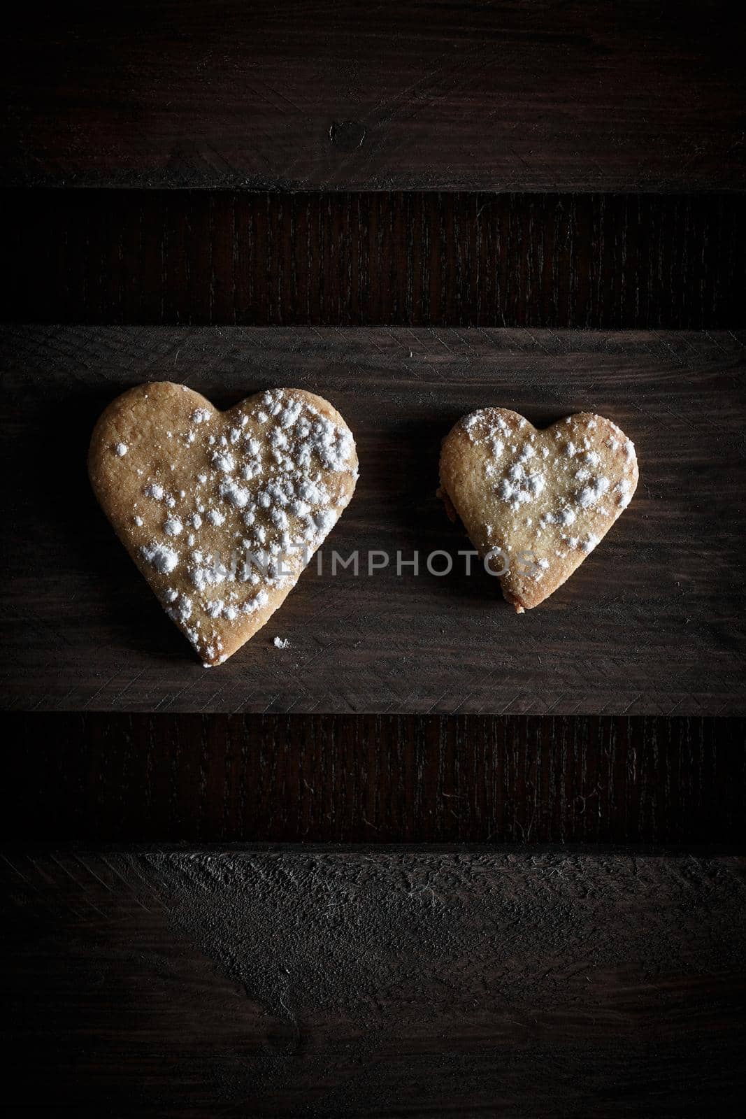 Two delicious home-made heart-shaped cookies sprinkled with icing sugar in a wooden board. Vertical image seen from above.Concept of love in couple. Dark moody style.