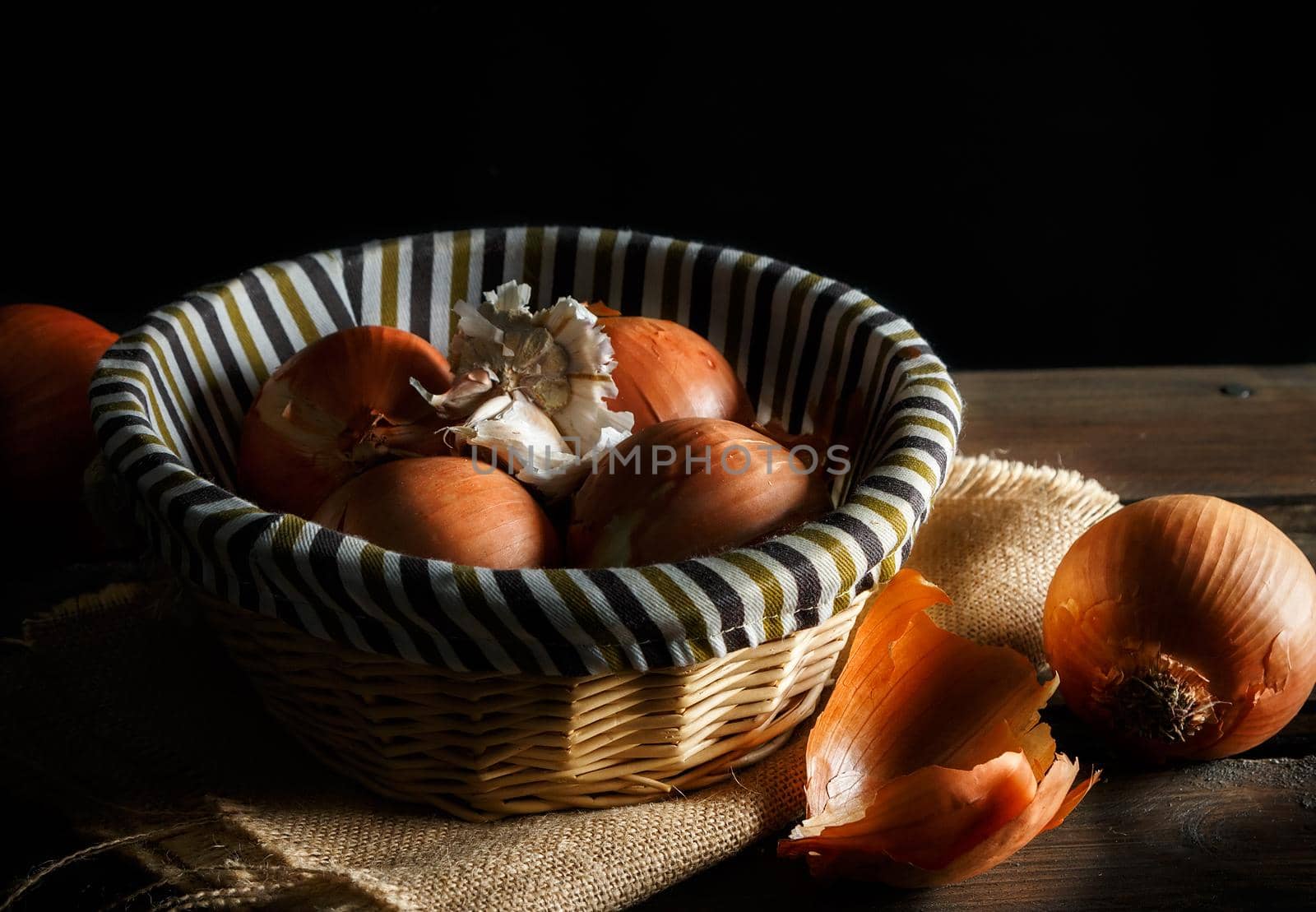 Onions and head of garlic in a wicker basket on wooden boards and sackcloth on black background. Rustic style. Horizontal image.