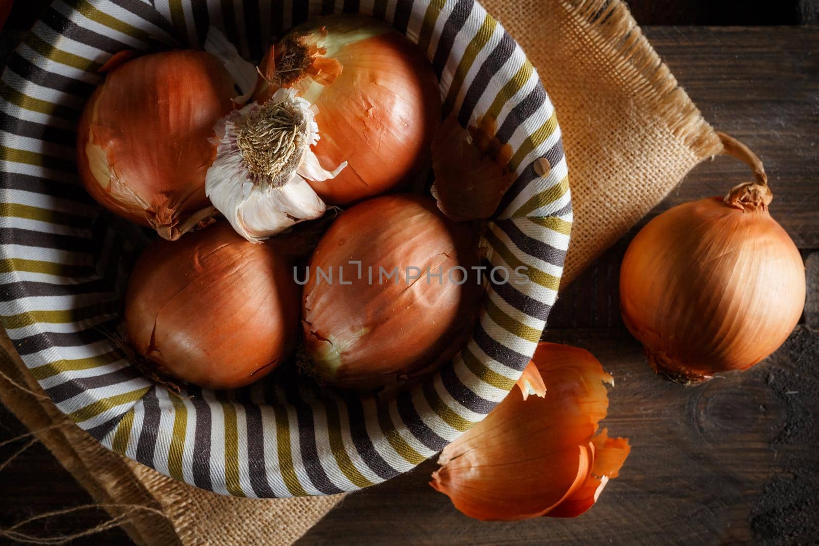 Still life of onions and garlic head in a wicker basket on a sackcloth and wooden boards. Seen from above. Rustic style. Horizontal image.