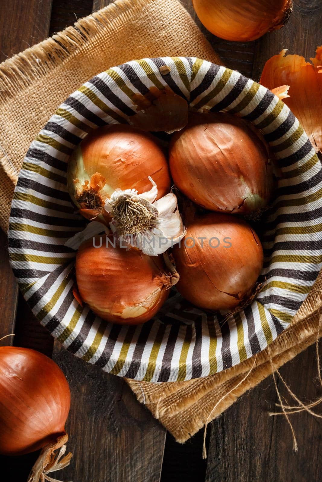 Still life of onions and garlic head in a wicker basket on a sackcloth and wooden boards. Seen from above. Rustic style. Vertical image.