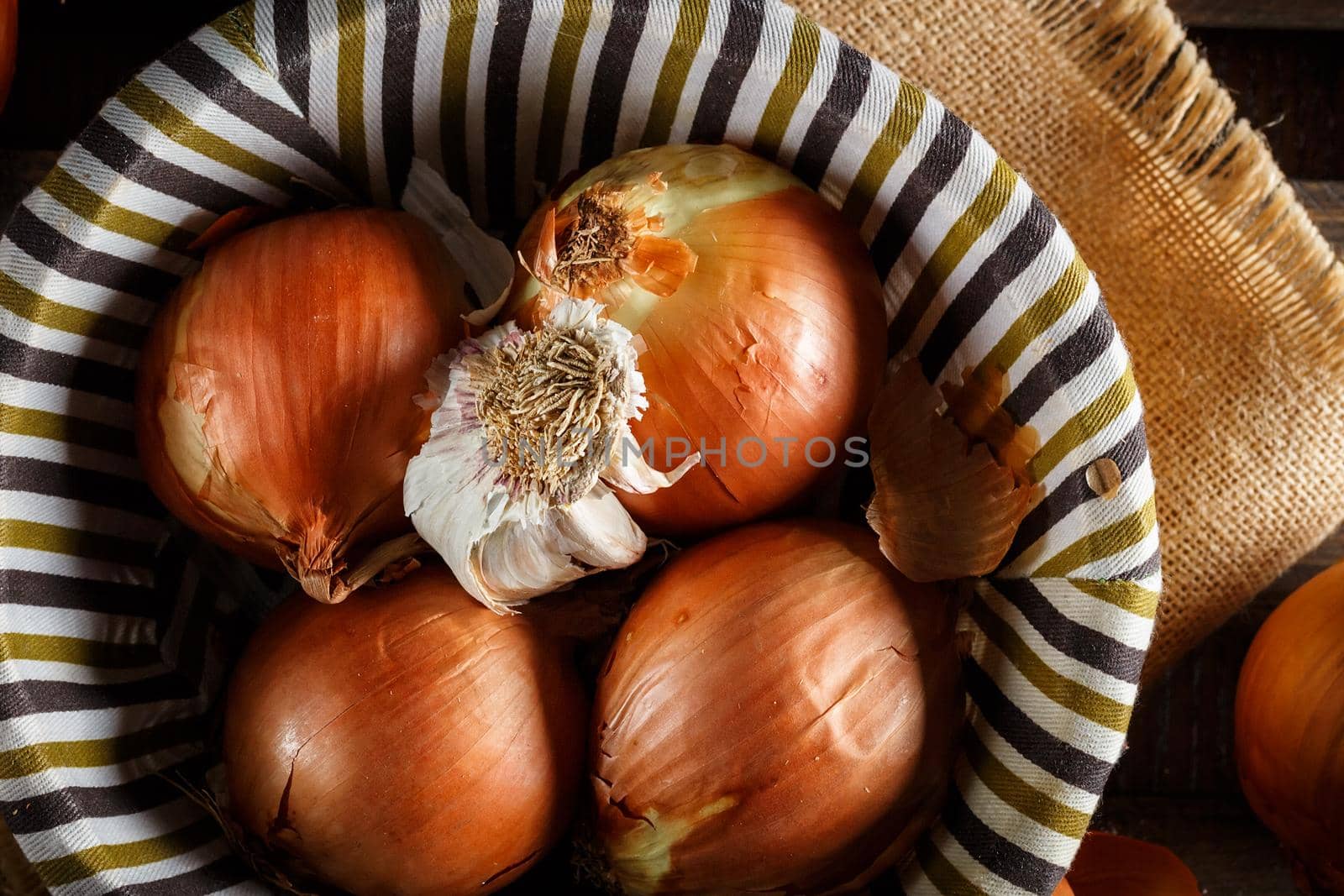 Still life of onions and garlic head in a wicker basket on a sackcloth and wooden boards. Seen from above. Rustic style. Horizontal image.