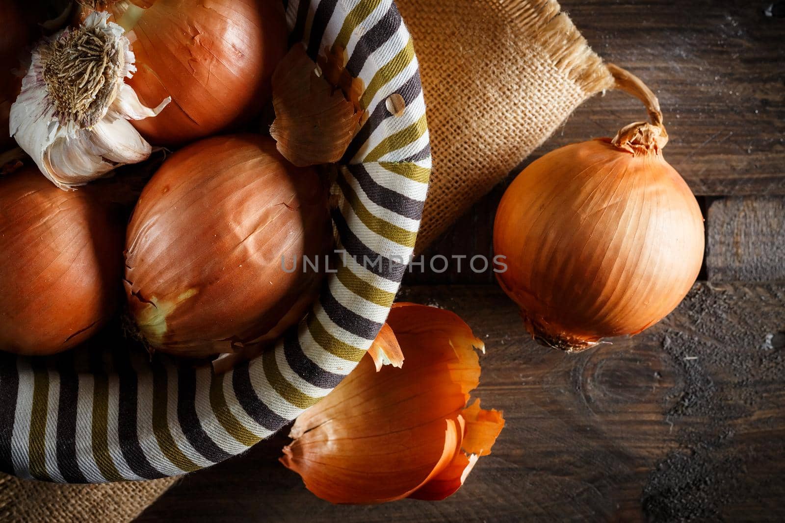 Still life of onions and garlic head in a wicker basket on a sackcloth and wooden boards. Seen from above. Rustic style. Horizontal image.