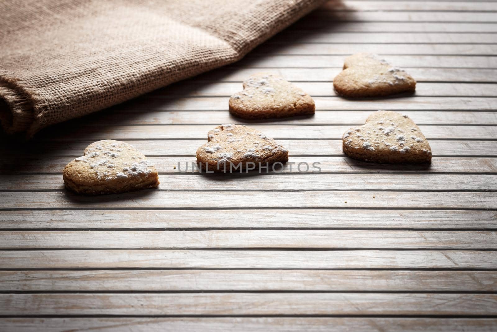 Delicious heart-shaped homemade cookies sprinkled with icing sugar on a wooden board with sackcloth in the background. Horizontal image seen against backlight.