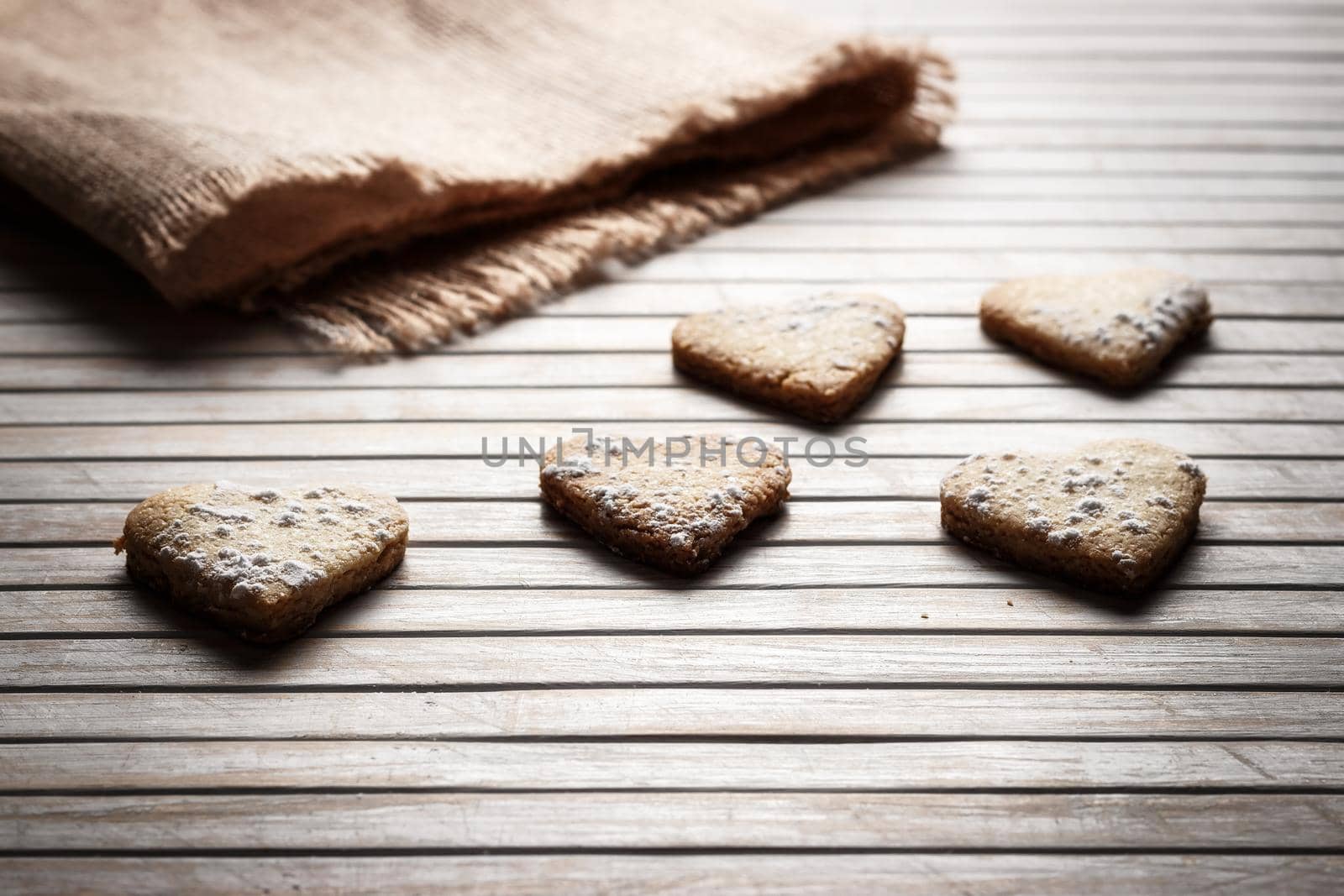 Delicious heart-shaped homemade cookies sprinkled with icing sugar on a wooden board with sackcloth in the background. Horizontal image seen against backlight.